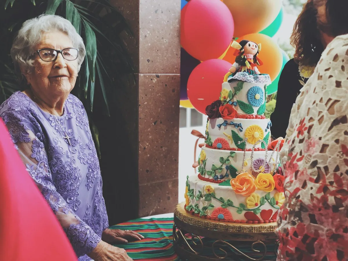 Grandmother beside a tiered birthday cake and balloons